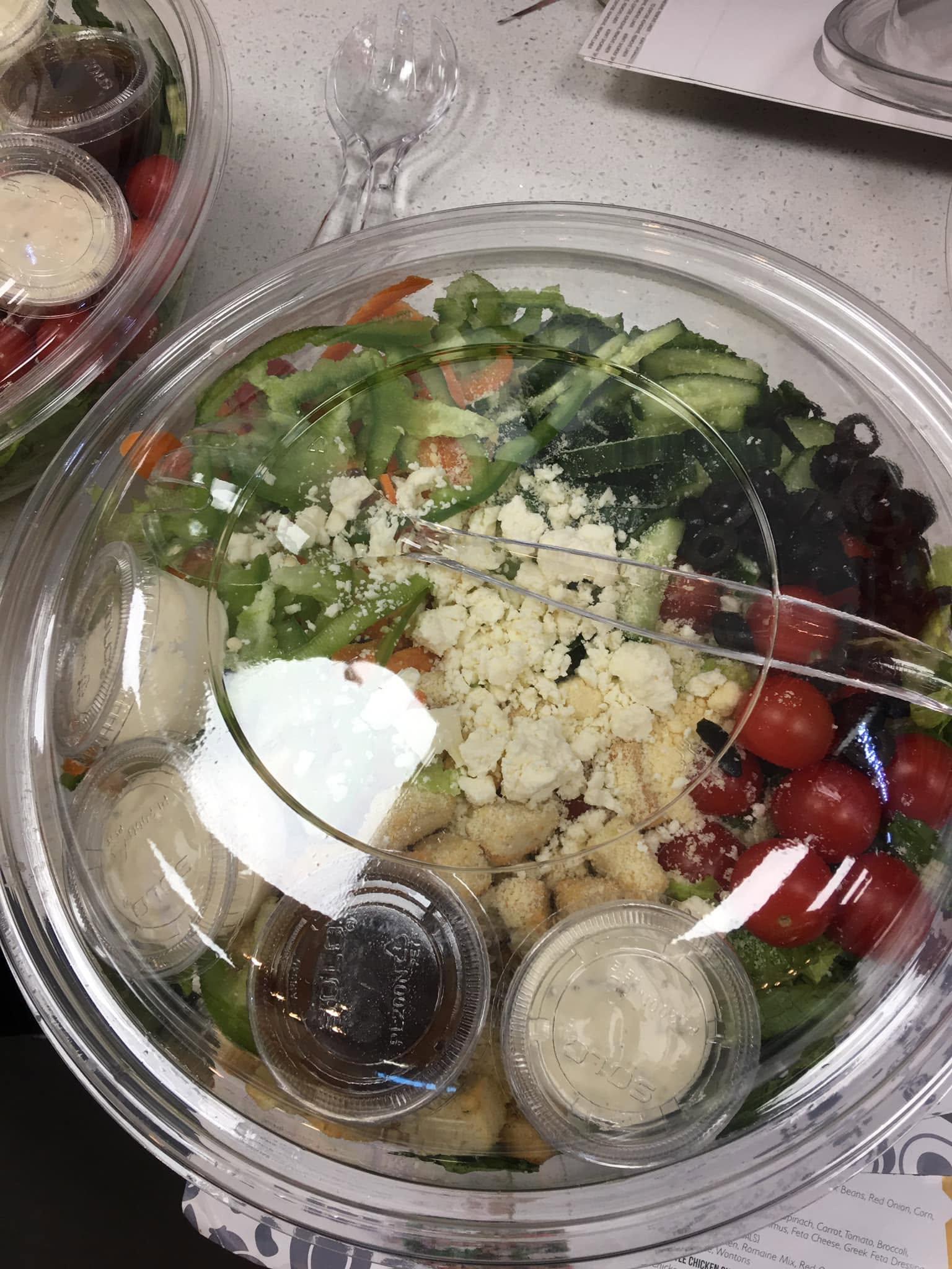 A large clear catering bowl of Greek salad at Fit For Life Cafe — romaine, green pepper, cucumber, carrot, cherry tomatoes, black olives, crumbled feta and croutons, with dressing cups tucked around the rim